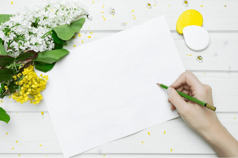 Hand about to write on blank piece of paper with floral accents on white table